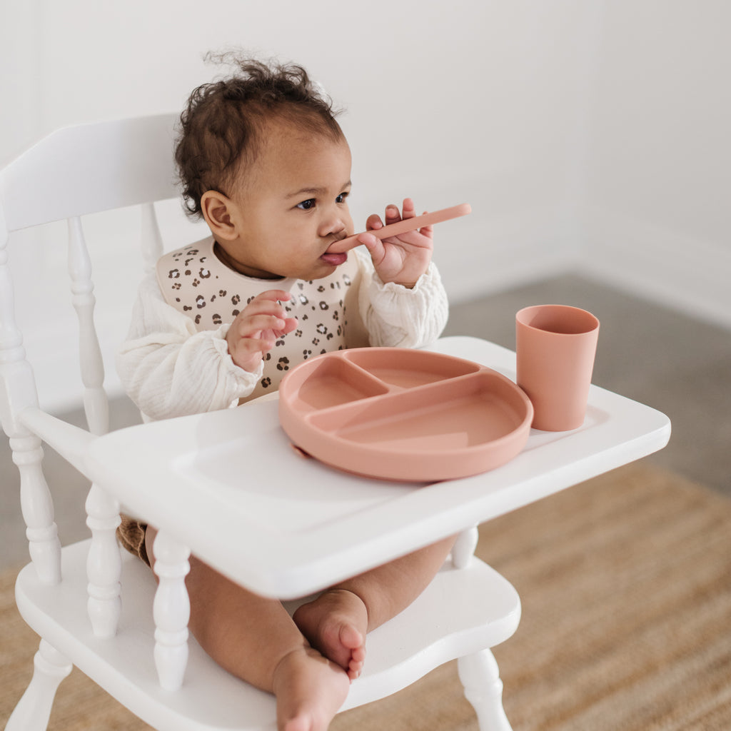 Silicone dinnerware set. MAde from 100% food grade silicone safety tested, free from BPA Phthalates and toxic metals. This image shows a blush toddler cup and toddler spoon with a slate blue three compartment divider plate