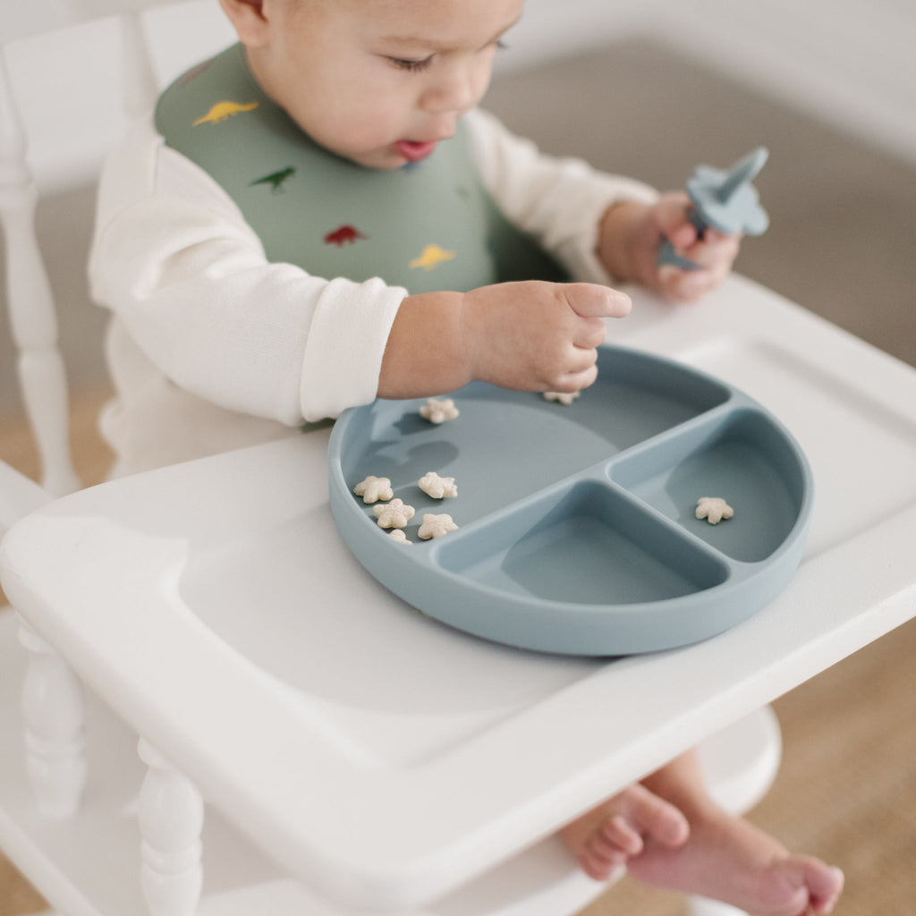 image of a little bowl holding a silicone training spoon eating from a pale blue silicone divider plate. he is wearing a sage green coloured dinosaur silicone bib