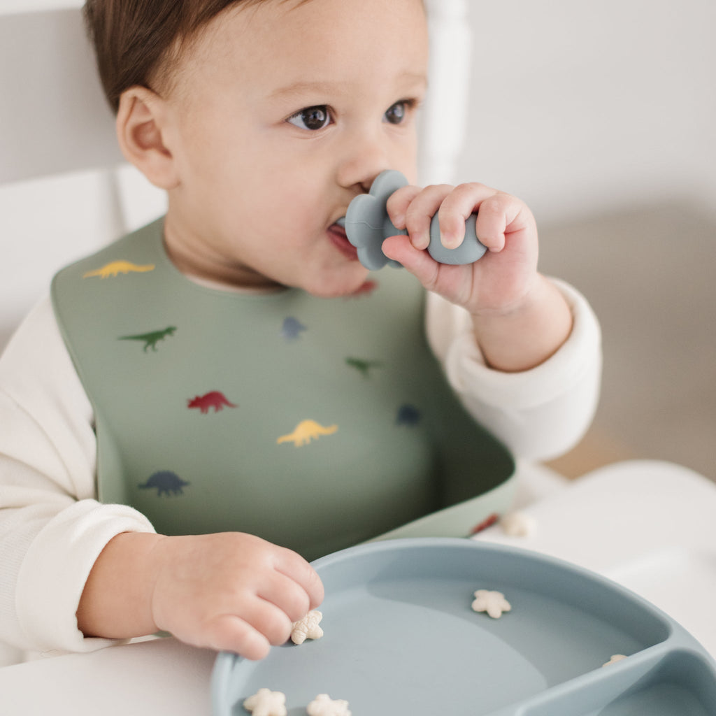 happy boy holds a training spoon in his mouth while wearing a sage green silicone bib with bright coloured dinosaurs printed on it