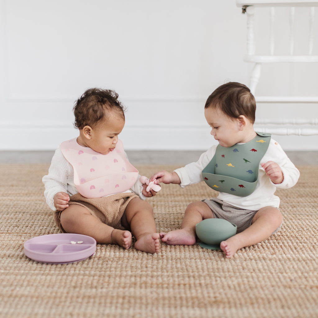 Two happy children sit eating food from a mauve divider plate and sage silicone bowl. The girl is wearing a blush rainbow silicone bib and the boy is wearing a sage dinosaur bib.