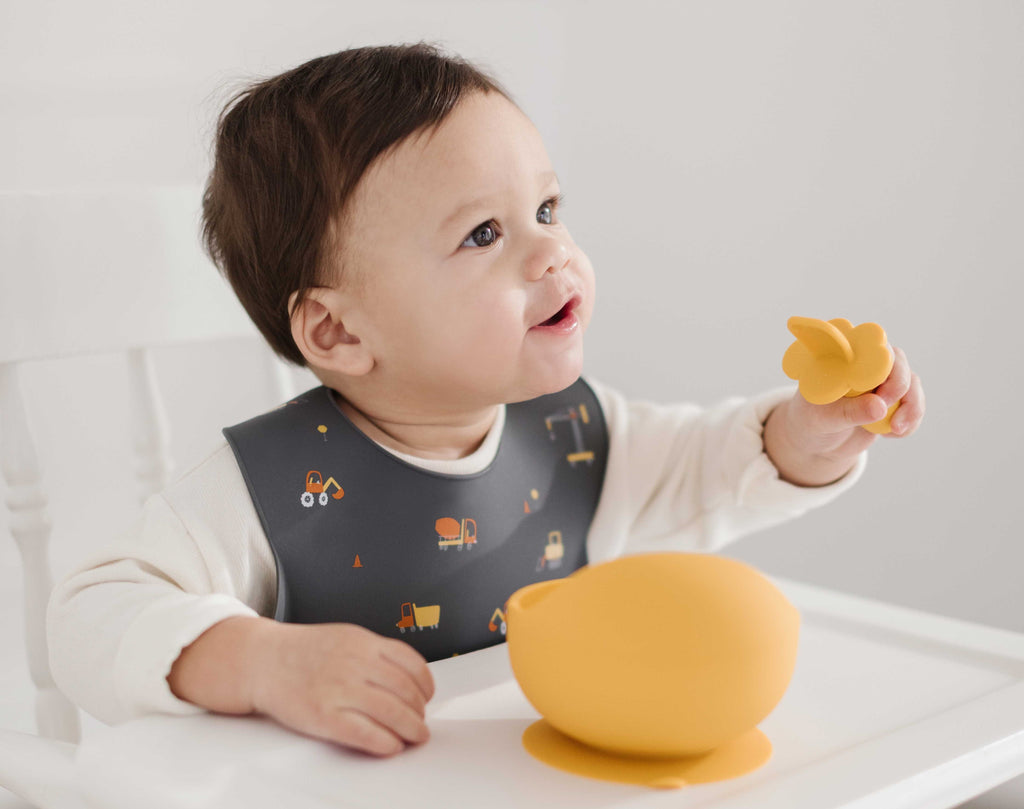 Little boy is sitting in high chair wearing lil north co construction silicone bib while using an infant training spoon and silicone bowl in mustard yellow to eat