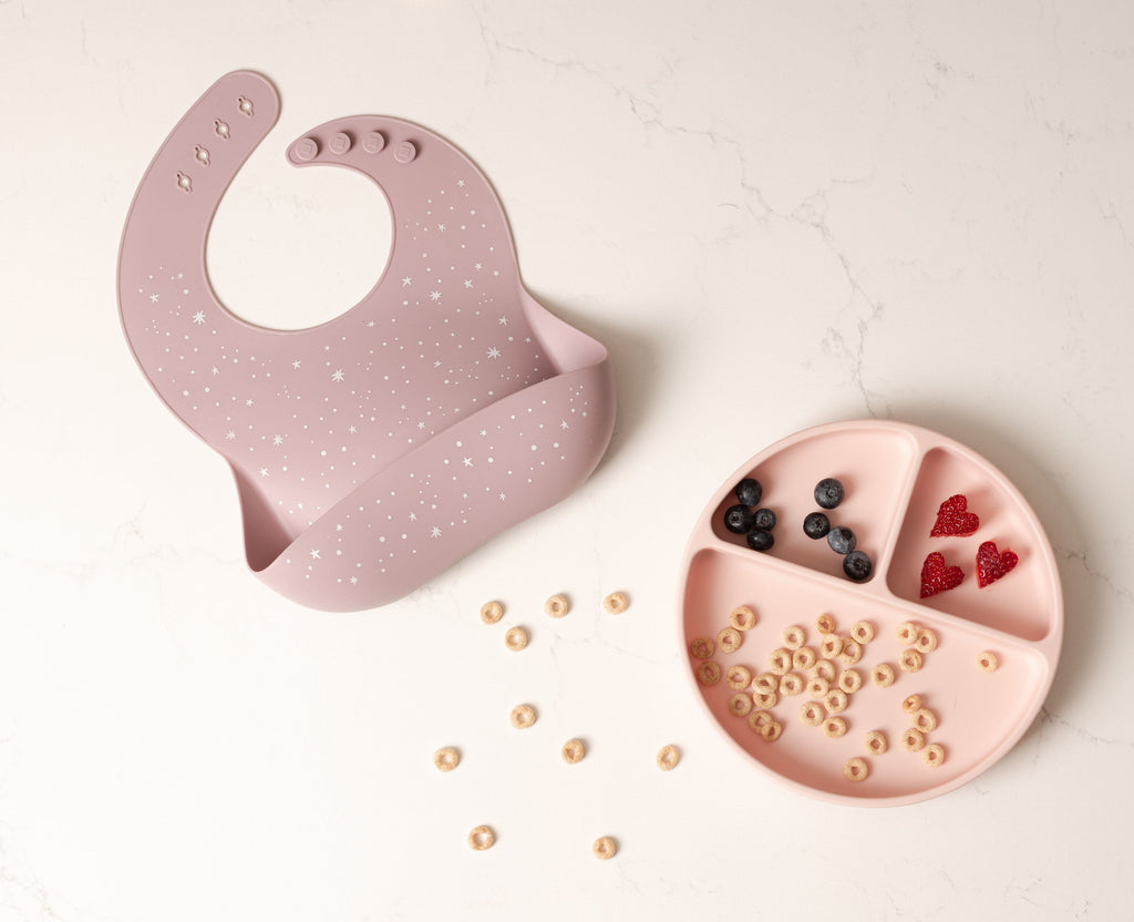 Mauve star silicone bib with deep front pocket lays on a counter next to a blush silicone divider plate holding cheerios blueberries and strawberries.