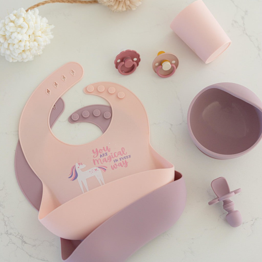 Two silicone bibs with large catch all pocket in blush and mauve lay on a white counter. A mauve bowl, infant training spoon, blush silicone cup and pacifiers sit beside it.