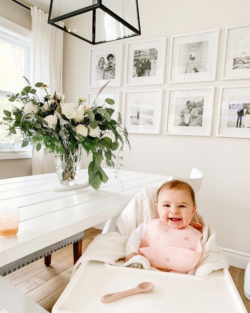 Happy baby sits in highchair wearing a silicone bib in blush with rainbows.