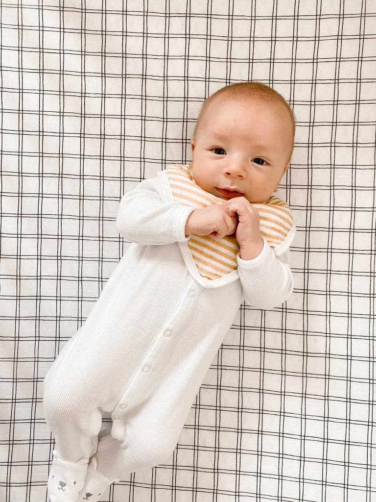 Image courtesy of @ginaraebarclay via instagram. Baby jack lays on a muslin grid crib sheet while smiling and wearing an ochre stripe muslin bib