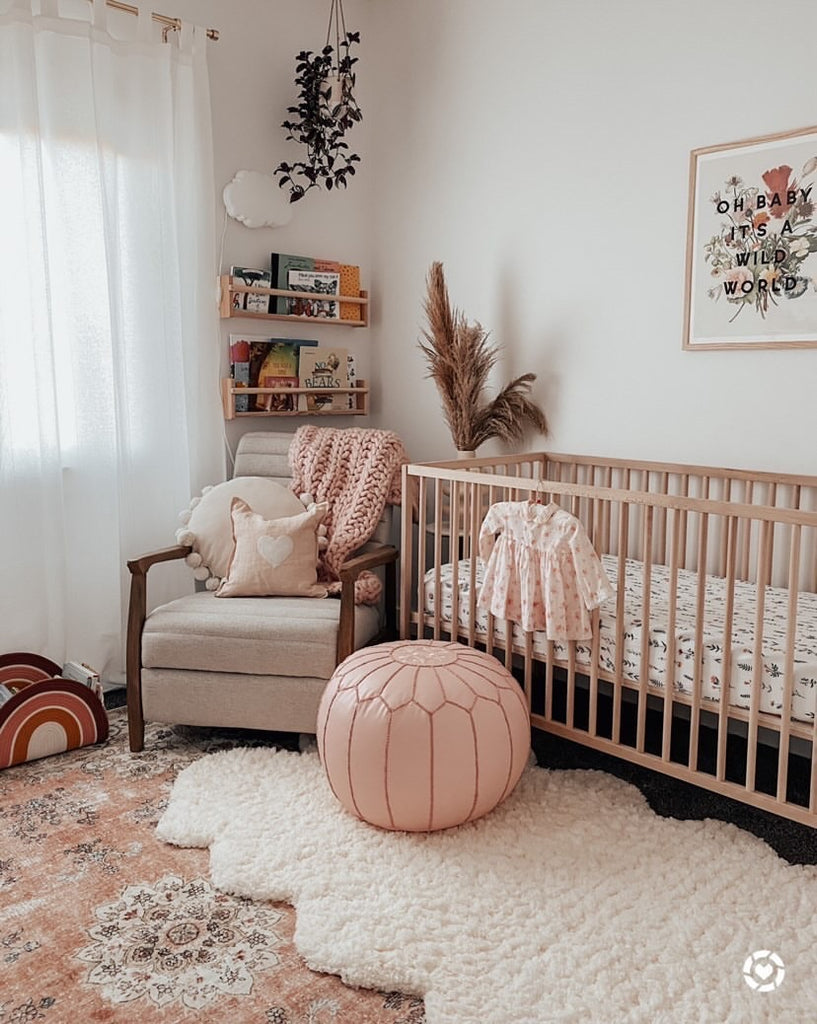 image courtesy of @theblushhome - Boho nursery featuring a natural crib with a wildflower patterned muslin crib sheet, blush pouf, beige glider and bookshelves