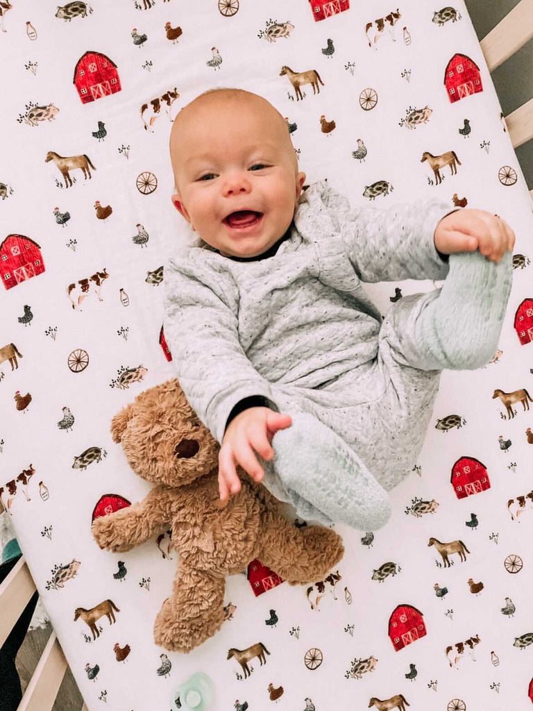 image courtesy of @lifeaftersped . Happy little boy lays on a farm animal crib sheet that includes a red barn. He is holding his feet in happy baby pose