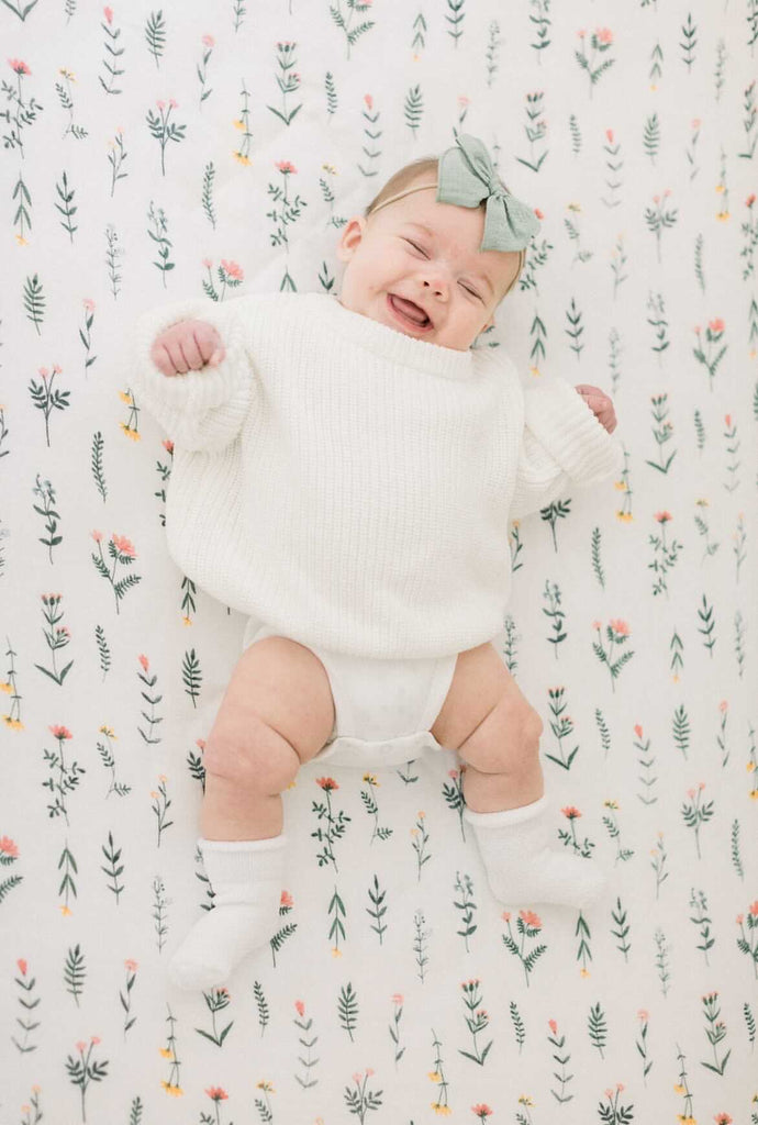 image of smiling baby girl in white sweater on a wildflower printed muslin crib sheet