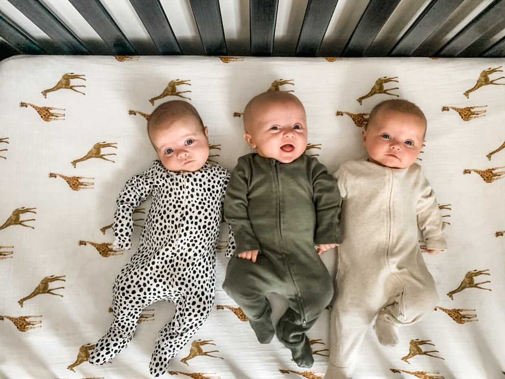 cute triplets lay on a giraffe printed crib sheet wearing neutral onesies.