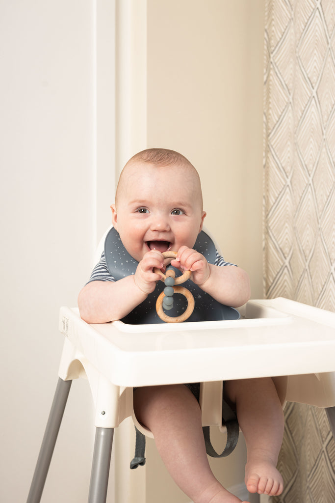 Happy baby sits in white high chair wearing a safety tested silicone bib in blue with tossed white stars.