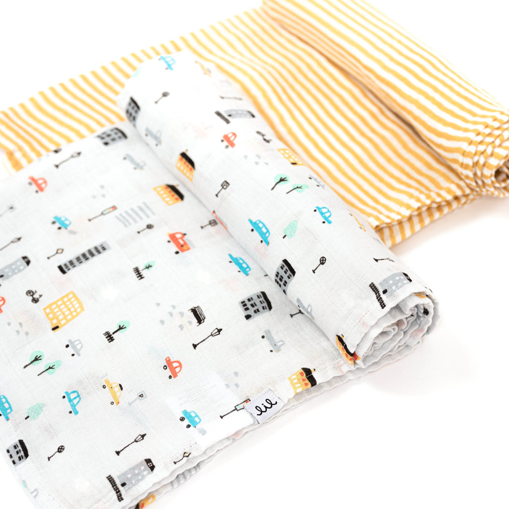 two swaddles rolled one blanket has a cute cityscape  print with grey ground and cars and trucks in yellow blurs and red grey buildings and street signs. Paired with ochre stripe swaddle blanket