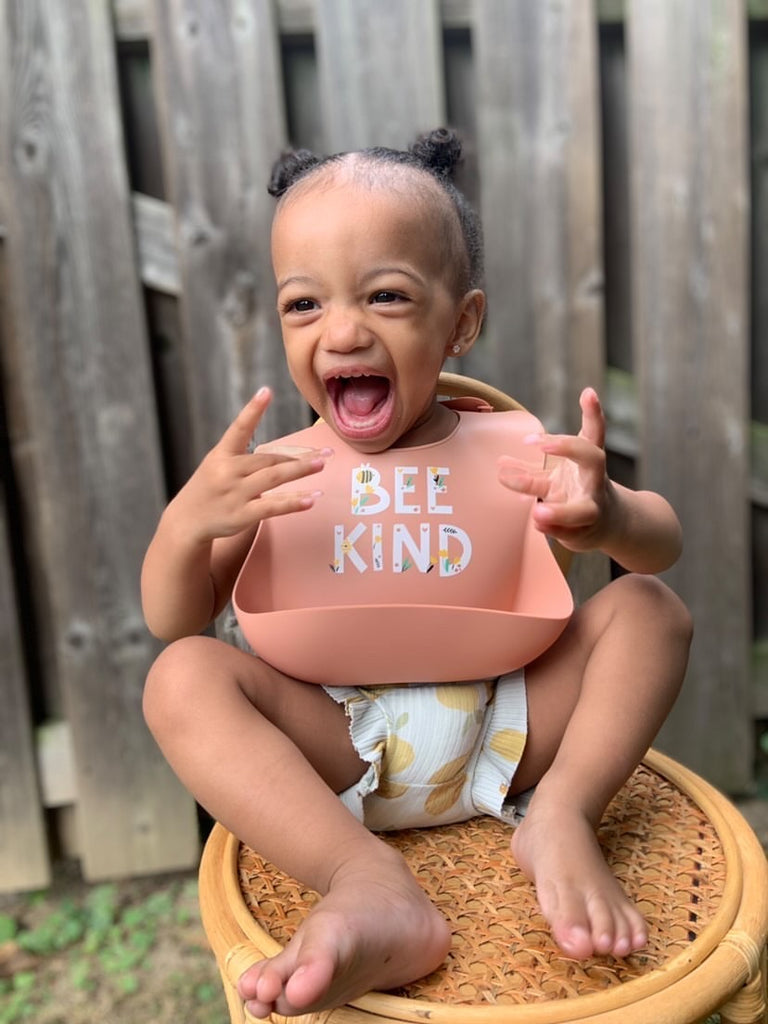 Happy little girl sitting on a chair wearing a terracotta coloured bib with the saying BEE KIND printed above the food catcher on the bottom.