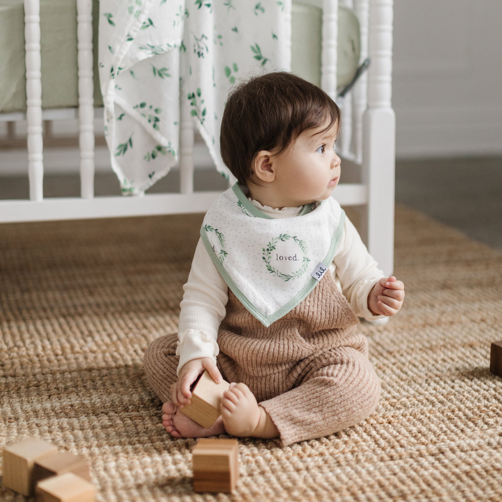 happy little girl sits on the floor playing with blocks while wearing a muslin bandana bib. the bib has a foliage wreath printed on it with loved syaing in the centre and has a sage green trim