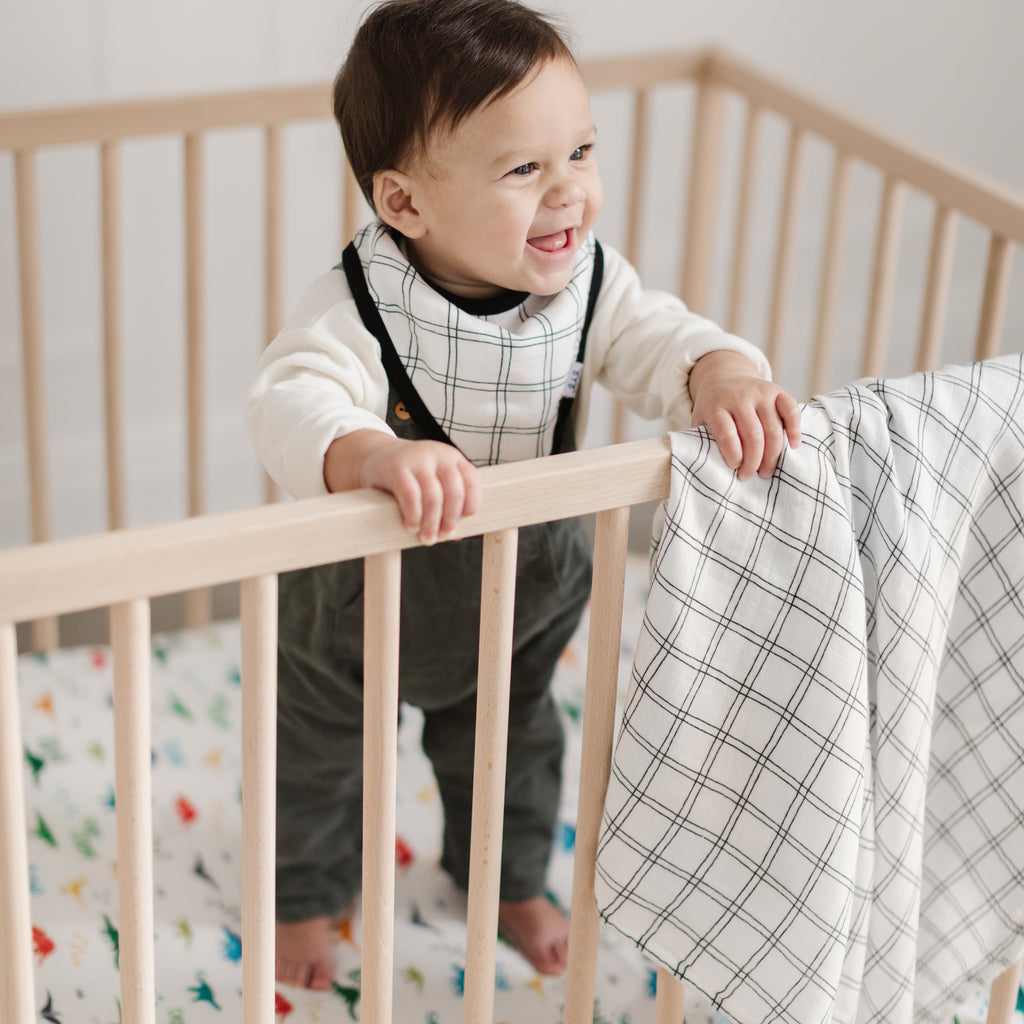 little boy happily stands in his crib while wearing a black and white grid patterned bandana bib. A matching swaddle hangs on the crib and a bright dino crib sheet is beneath him
