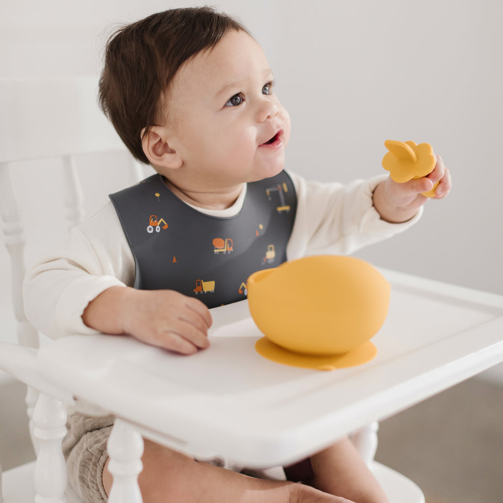 little boy wearing dark grey silicone bib with construction trucks printed on the bib.  Little bowl is wearing the bib while eating from a mustard bowl with an infant training spoon made from food grade silicone