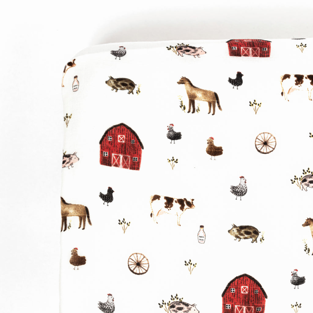 Corner image of premium bamboo cotton muslin crib sheet featuring farm animal print that has red barn and various farm animals 