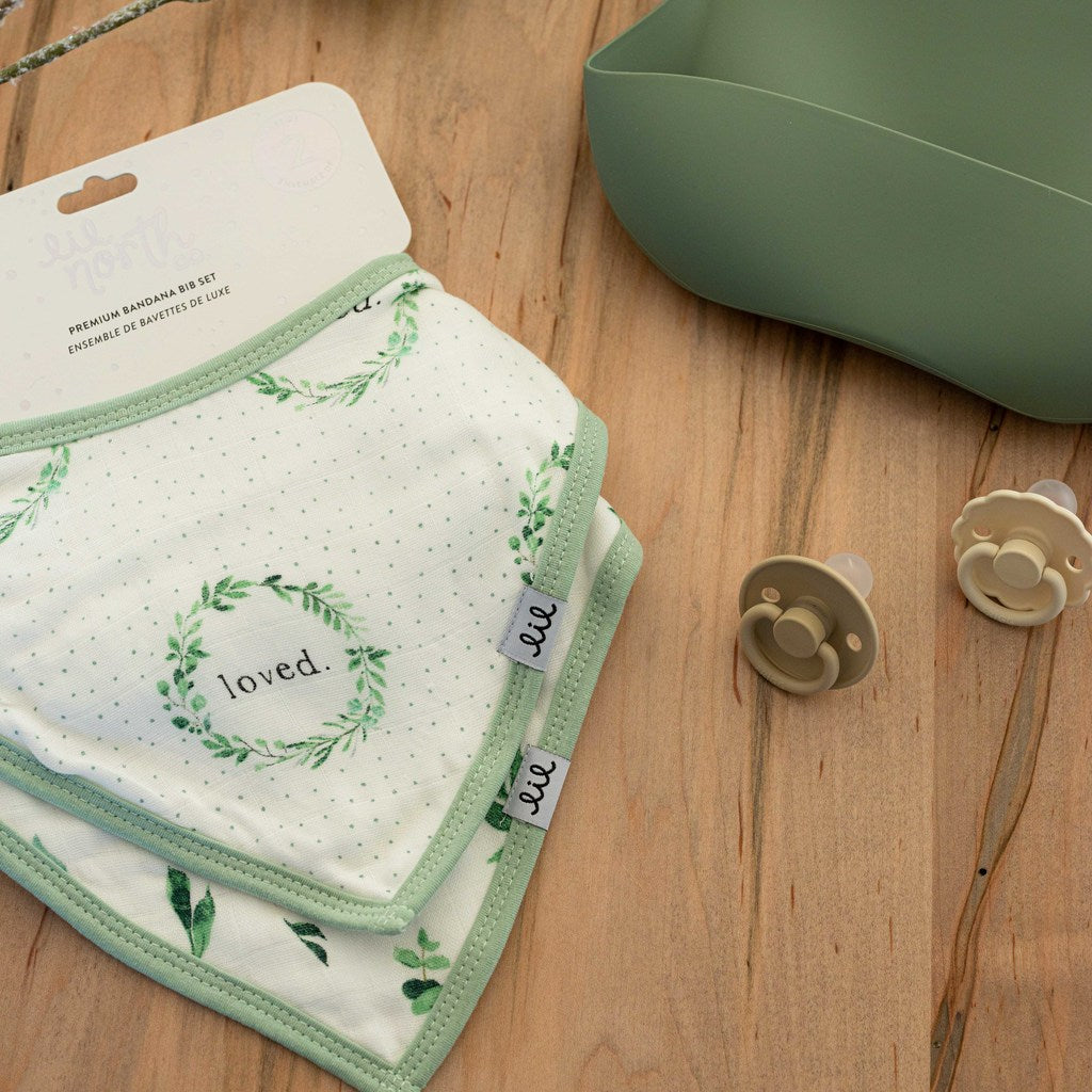 packaged muslin bibs sit on a wood table. The bibs have a sage green trim. One is printed with a foliage wreath that says loved in the middle and the other has a tossed green foliage print on it.