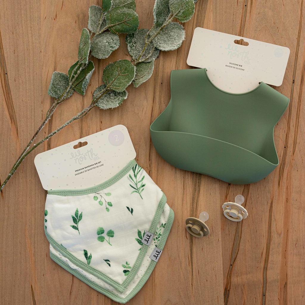 image of sage silicone bib and a 2 pack of bandana bibs in watercolour foliage print laying on a wood table