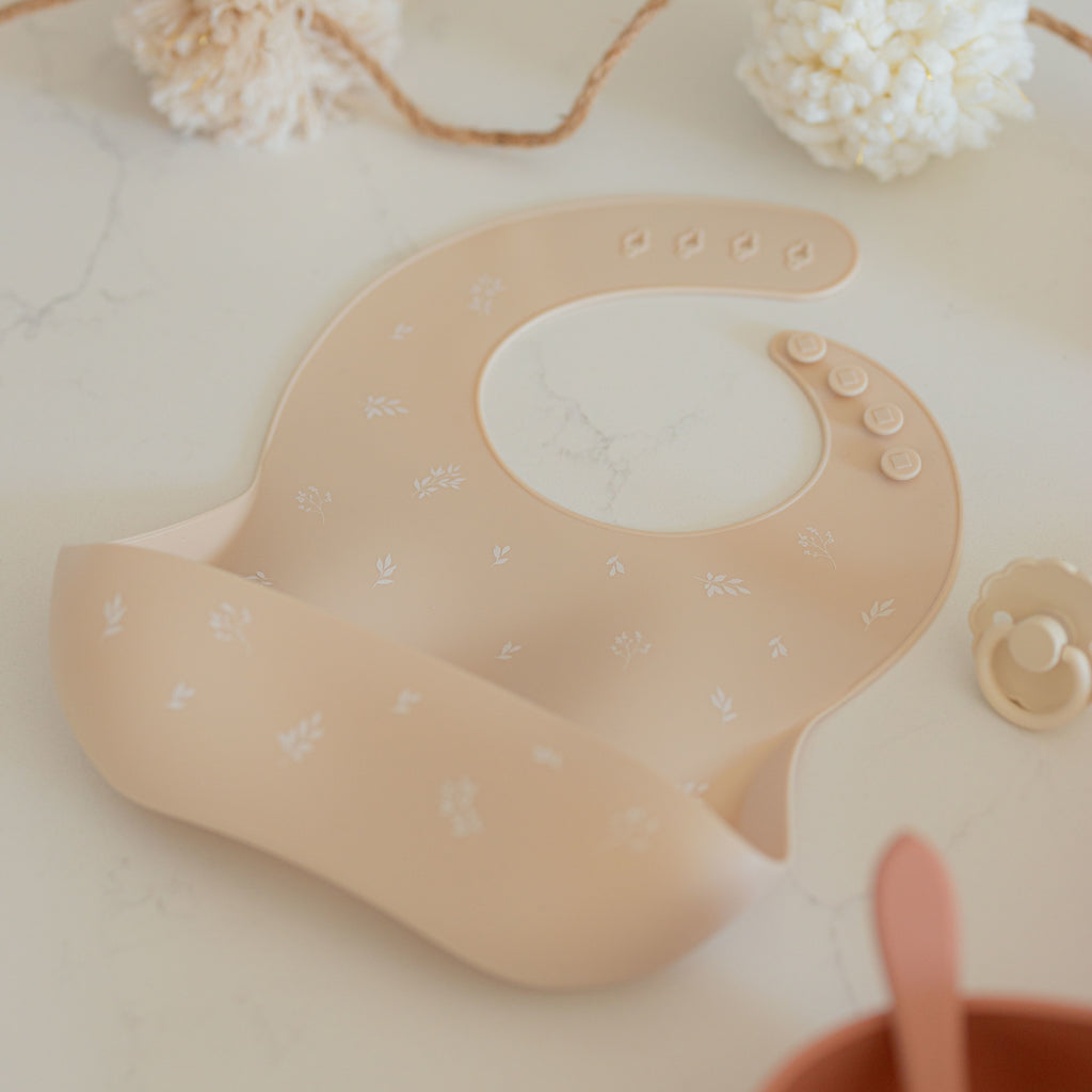 Closeup image of silicone bib on a counter. The bib is a sand colour with white foliage printed on it.