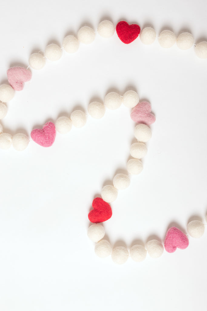 image of a valentines felt garland for decor. the garland includes white felt balls and felt hearts in red and shades of pink. There are 5 white balls between each heart.