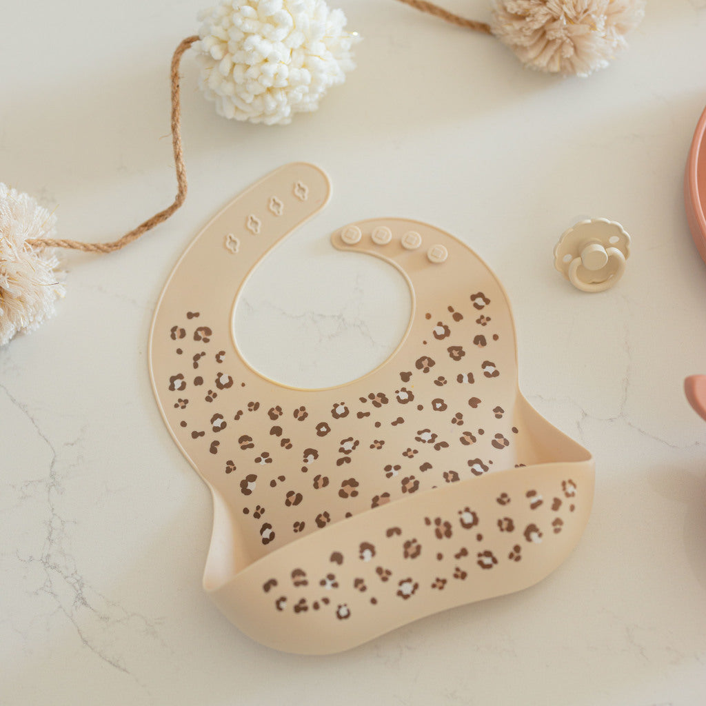 Cheetah silicone bib lays on a counter. Bib has deep front pocket and four adjustable neck strap settings.