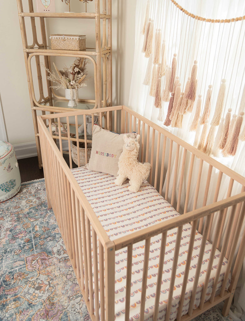 Lifestyle image of beautiful boho nursery featuring ikea sinklar crib vintage rattan bookshelf styles with lil north co linear rainbow crib sheets. Accessorized with homebody pillow and llama stuffed animal