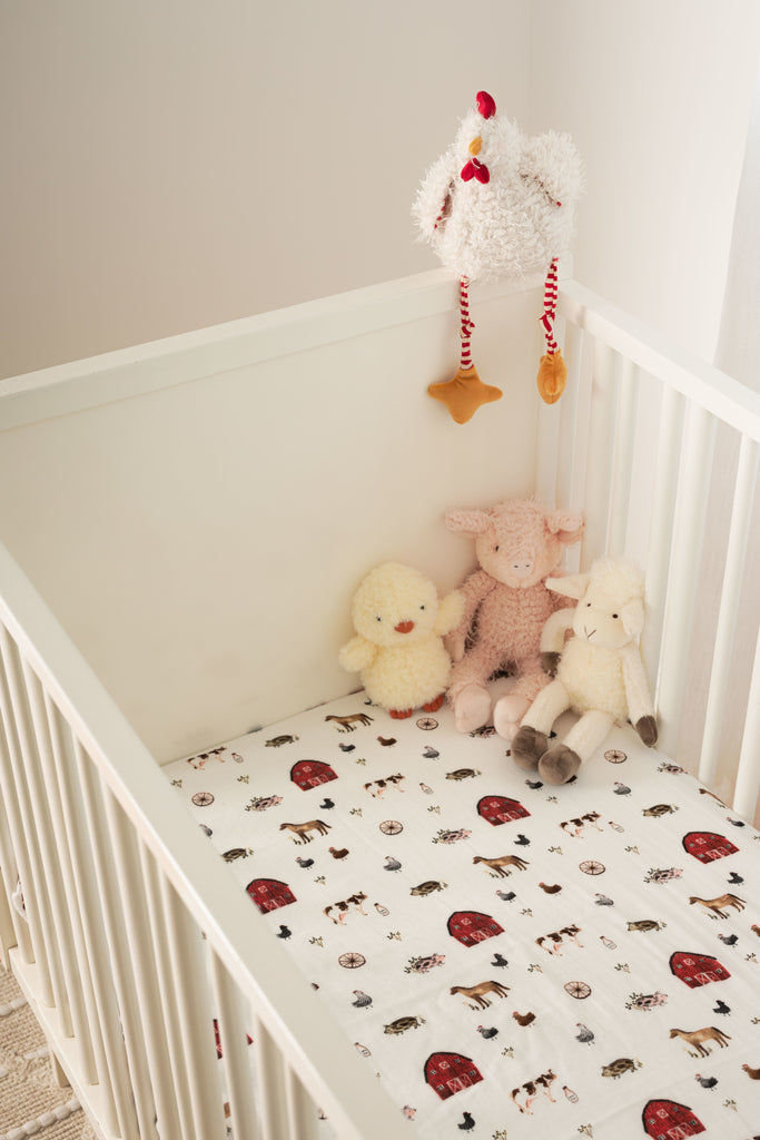 Lifestyle shot of farm animal muslin crib sheet in a white crib. Stuffed arm animal dolls sit in the corner of the crib.