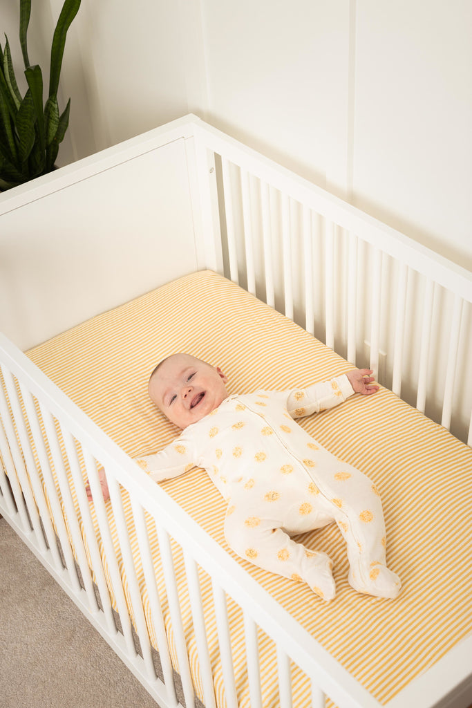 happy baby in sunshine sleeper lays in white crib on top of ochre and white stripe bamboo cotton muslin crib sheet