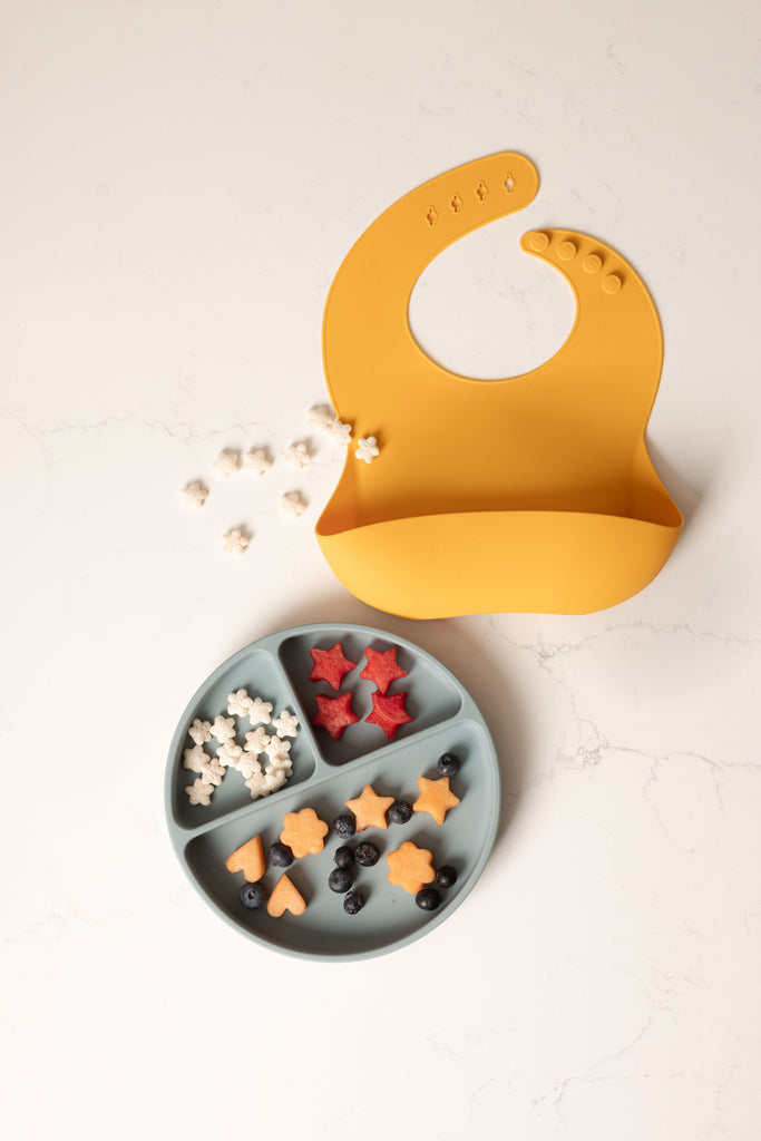 image of a mustard silicone bib with large catch all pocket lays on a counter. Beside the bib is a pale blue divider plate