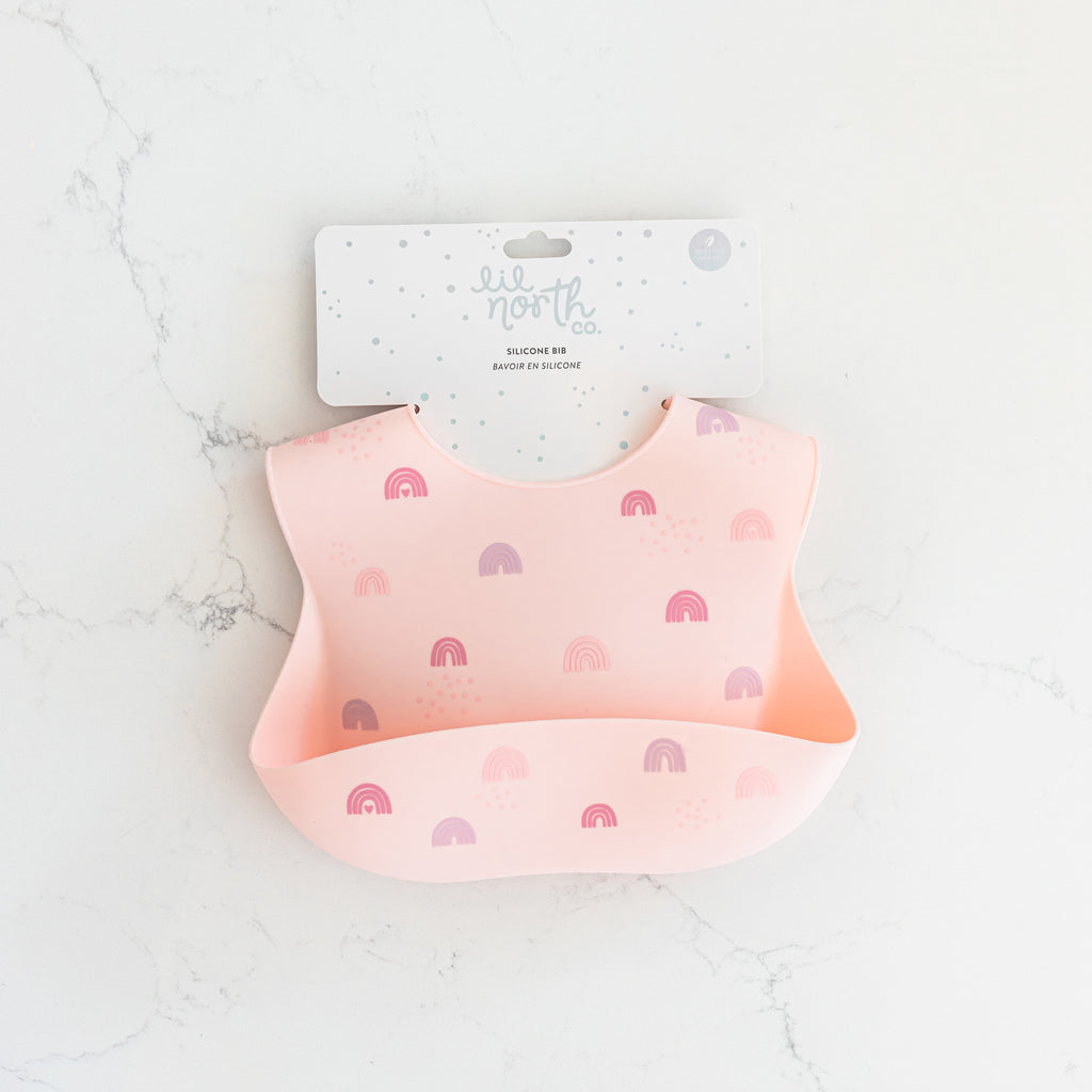 Safety tested silicone bib in blush with tossed rainbows in pink and purple tones.