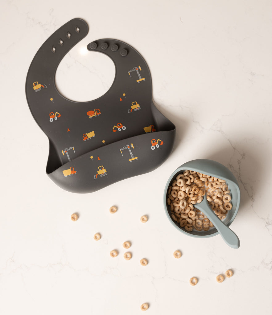 Silicone bib in charcoal grey with small construction trucks printed on it in yellows and oranges. Beside the bib is a pale blue silicone bowl and spoon filled with cheerios and milk.