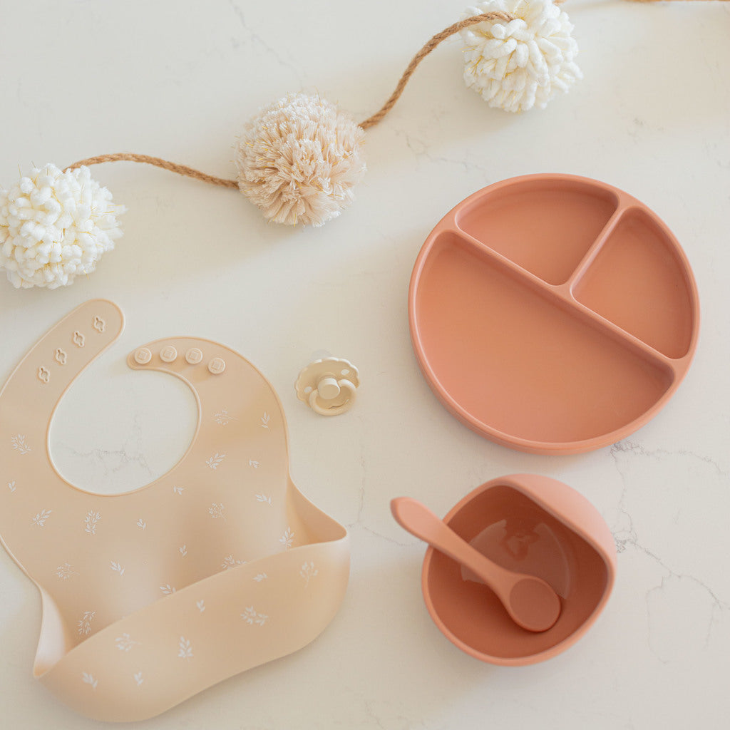 Silicone bib sits on the counter. The bib is a sand colour with white foliage printed on it. Beside the bib is a terracotta divider plate, bowl and spoon all made from food grade silicone to help babies transition to solids.