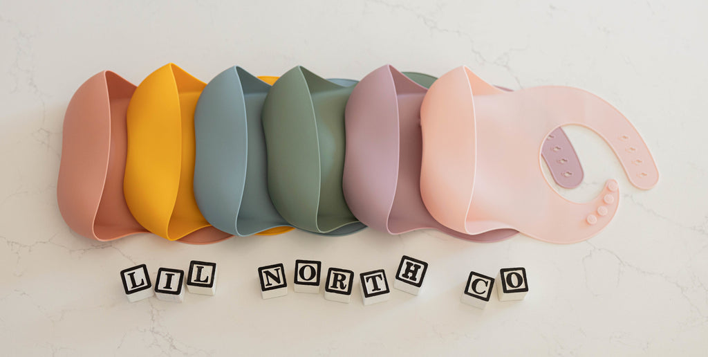 6 solid coloured silicone bibs lay on a white counter. The bibs are in terracotta, mustard, pale blue, sage, pale mauve and blush and feature a large front pocket to catch food.