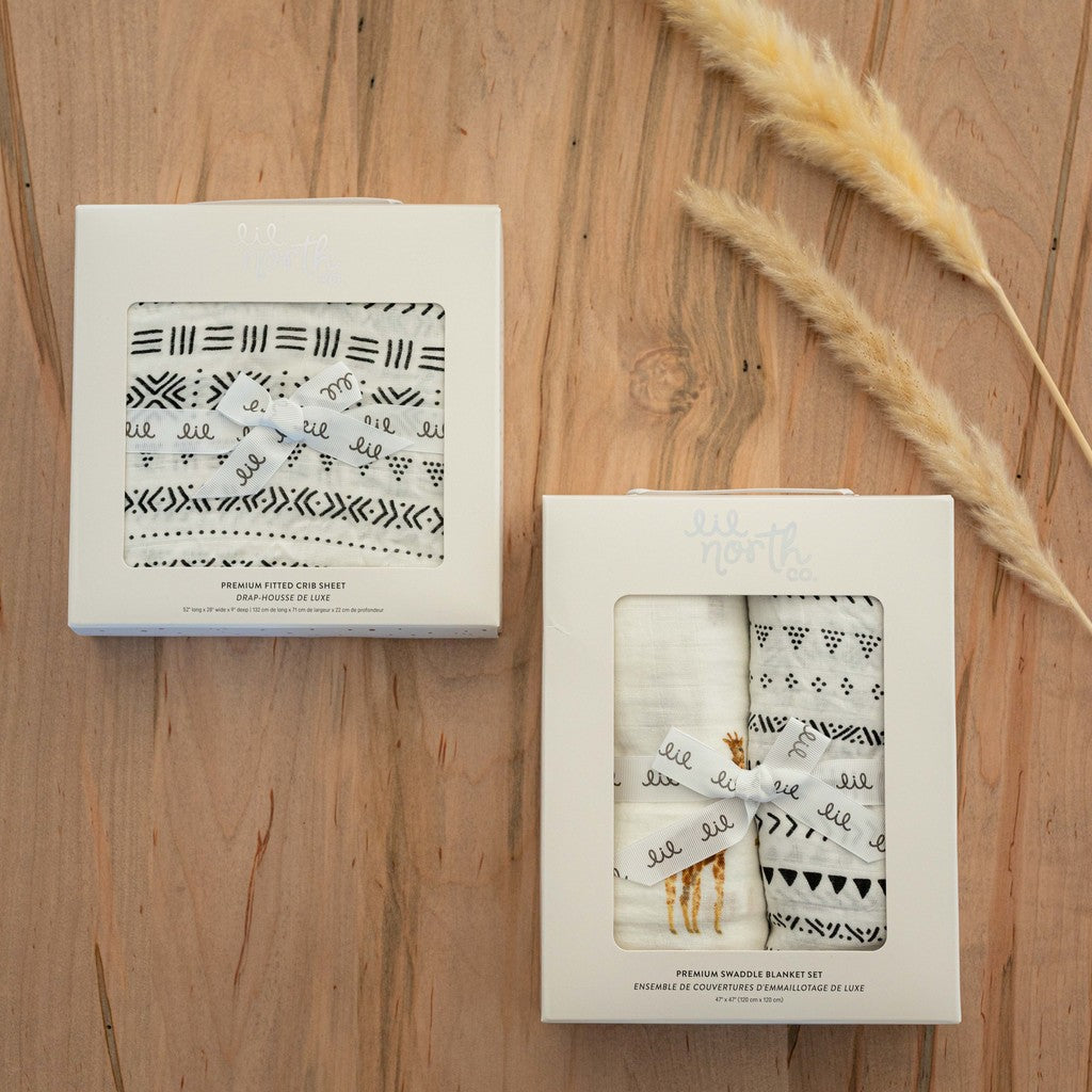 image of packaged product on a wood table. the crib sheet is in a white box and is a black and white tribal pattern. the swaddle set is in a white box with a giraffe swaddle and a black and white tribal pattern