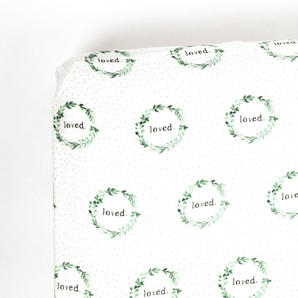 Corner image of premium bamboo cotton muslin crib sheet. Print features watercolour foliage wreath with typewriter font loved saying in the middle. Sage dots scatter in the background of the print.