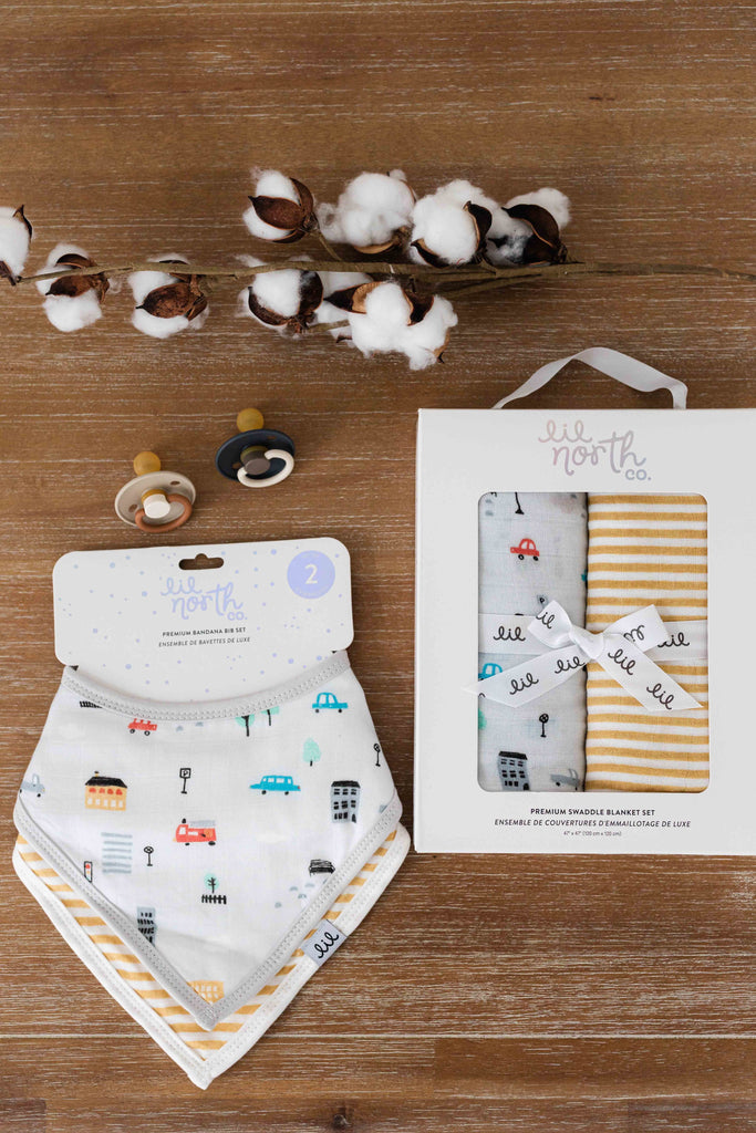 a baby gift assortment lays on a wood table. the set includes a two pack of muslin swaddles and a two pack bandana bibs. The printed are a city cars sketch in grey and some pop colours and an ochre stripe pattern.