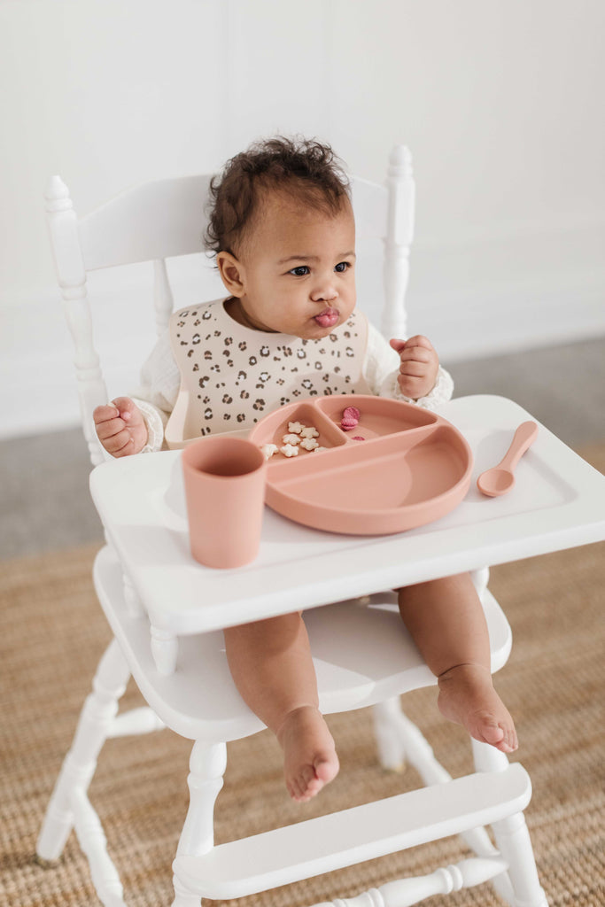 Little girl sits in highchair while eating. She is wearing a cheetah print silicone bib using a terracotta divider plate, cup and spoon made from food grade silicone.