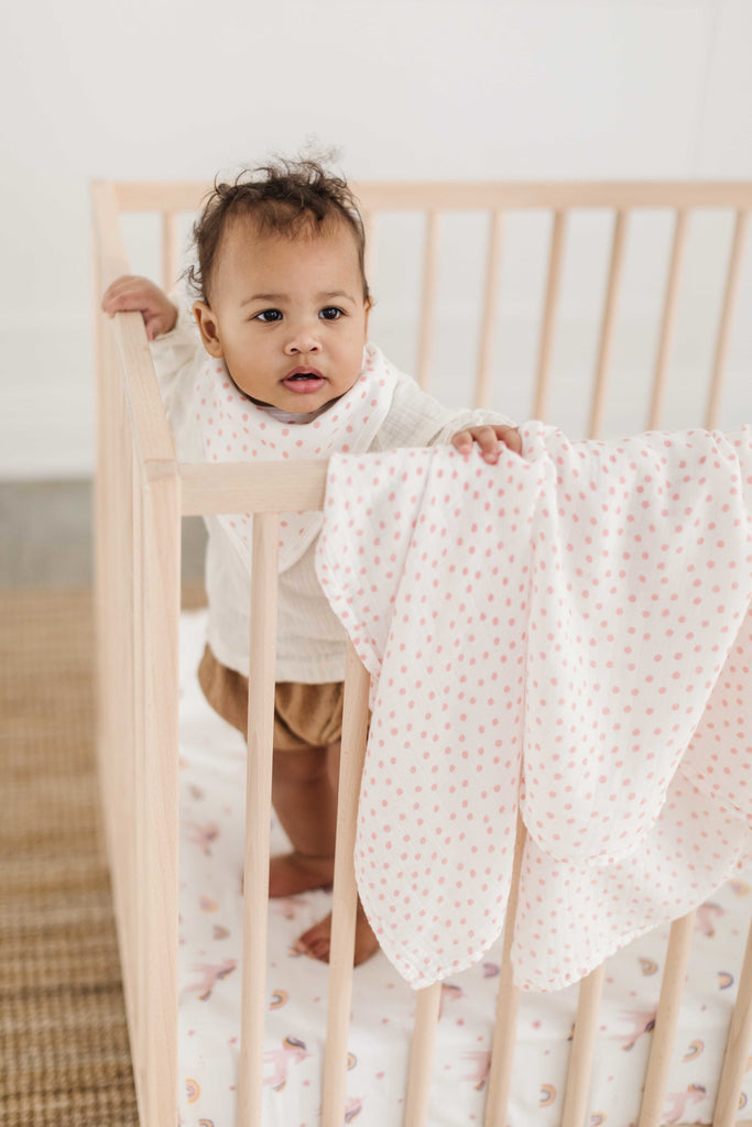 image of little girl standing in a natural wood crib. she wears a blush dot muslin bib and holds a matching swaddle.