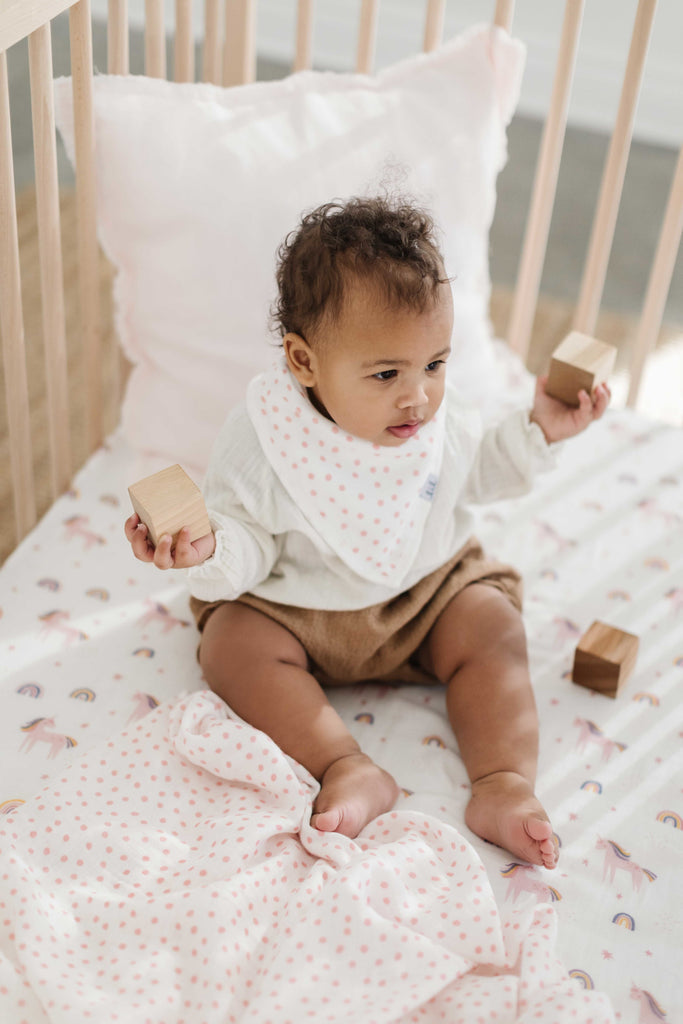 little girl sits in a crib while playing with blocks. She is wearing a blush dot printed bandana bib and sitting on a pastel coloured unicorn crib sheet. Behind her is a blush linen decorative pillow.