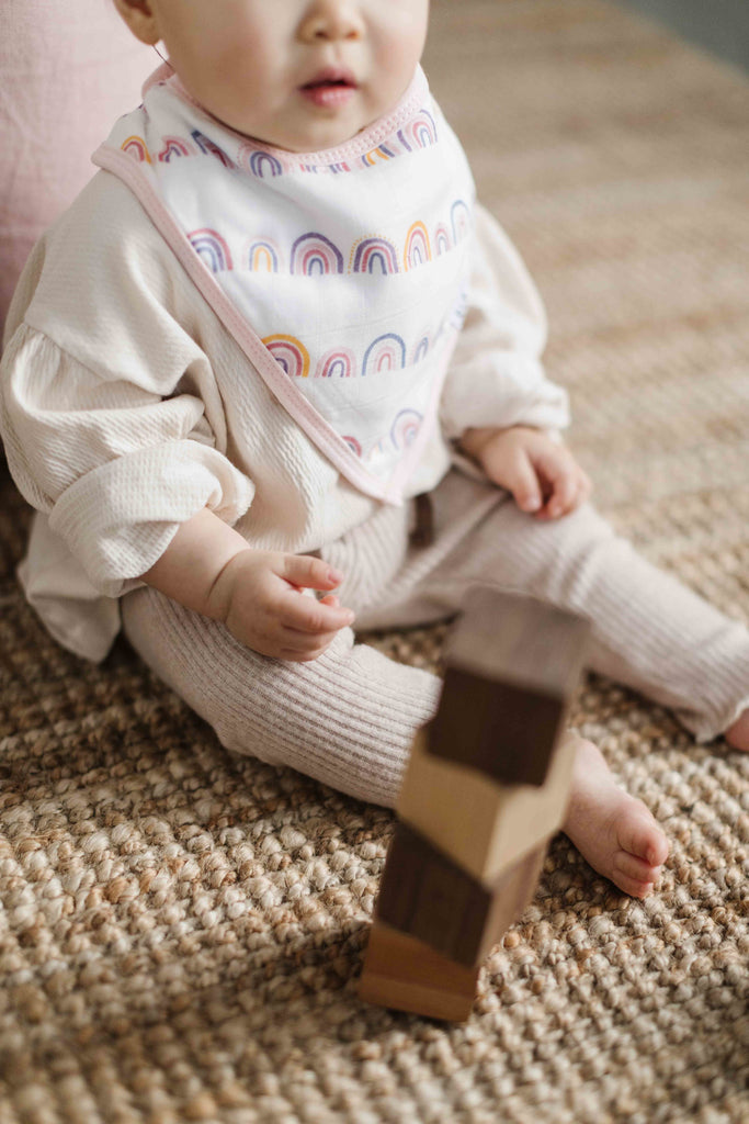 a little girl sits beside blocks wearing a rainbow printed muslin bandana bib with pink trim