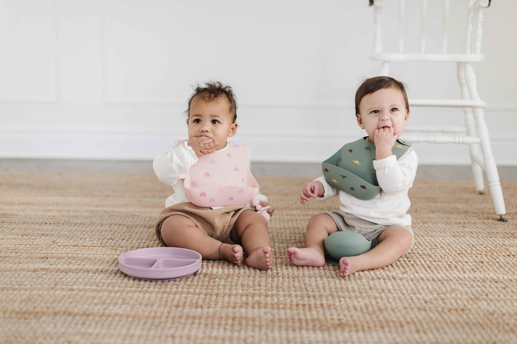 Two small children sit on the floor eating cheerios. The boy is wearing a silicone dinosaur bib in green and holding a matching silicone bowl. The little girl is wearing a pink rainbow silicone bib and eating from a pale mauve silicone divider plate.