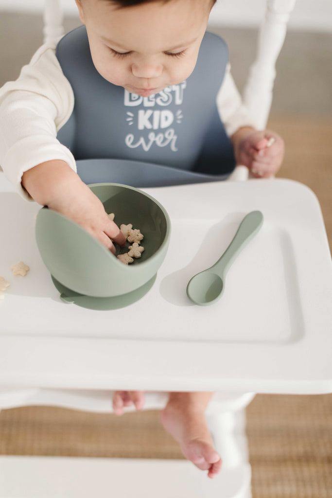 Little boy reaches into a sage green bowl filled with star puffs. He is wearing a slate blue silicone bib that says best kid ever in fun white writing. A sage green silicone spoon rests on his food tray.
