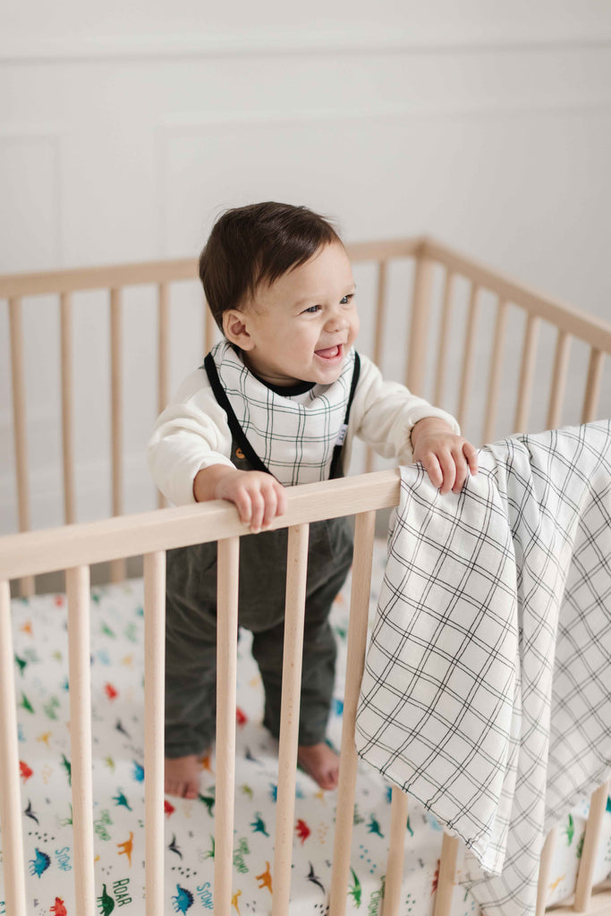 happy boy stands in crib while wearing a black and white grid bandana bib, a matching swaddle hangs on the crib. Beneath his feet he stands on a bright dino crib sheet
