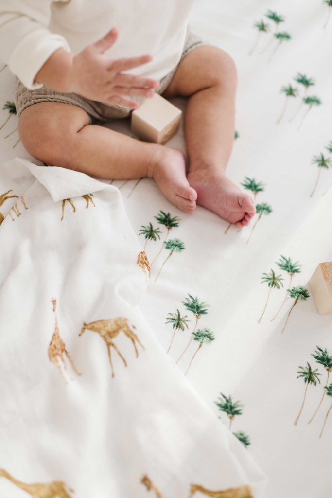 little feet sitting on a palm tree printed muslin crib sheet. He is holding a watercolour giraffe swaddle