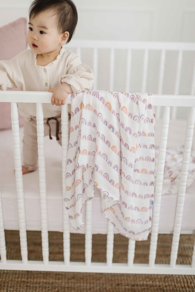 image of little girl standing in a white crib. In the crib is a blush linen crib sheet, a dusty rose linen pillow. Hanging from the crib is a rainbow patterned muslin swaddle.