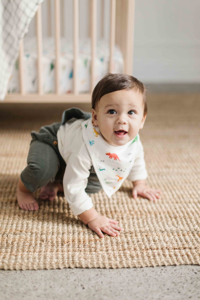 little boy crawls on the floor while wearing a bright dino printed muslin bandana bib