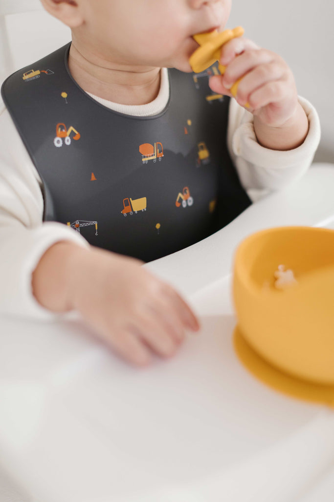 Close up image of dark grey silicone bib with construction trucks printed on the bib.  Little boy is wearing the bib while eating from a mustard bowl with an infant training spoon made from food grade silicone.