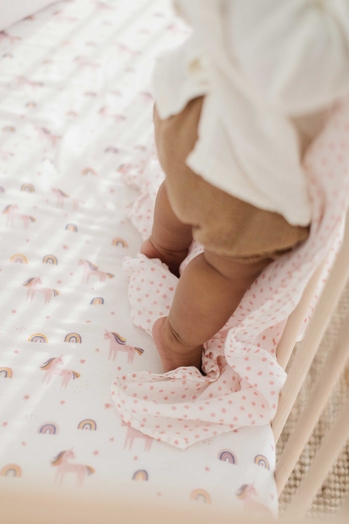 close up image of little feet standing in a crib while holding a blush dot muslin swaddle and standing on a unicorn rainbow printed crib sheet