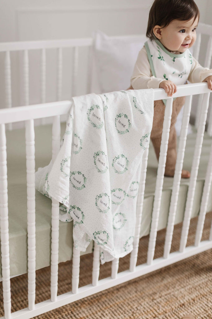 image of happy girl standing in white crib. inside the crib is a sage linen crib sheet, a white linen decorative pillow. Hanging on the crib is a loved wreath foliage swaddle.