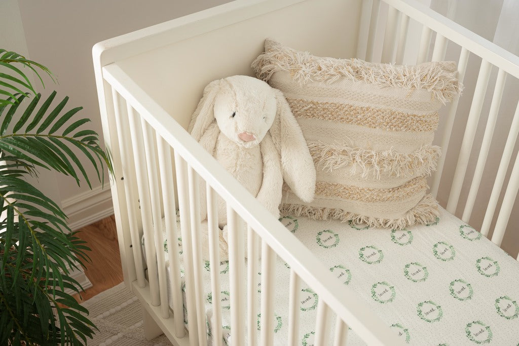 Image of a white crib. In the crib is a muslin crib sheet with a soft green foliage wreath that has loved printed inside it. inside the crib is a pillow and stuffed animal